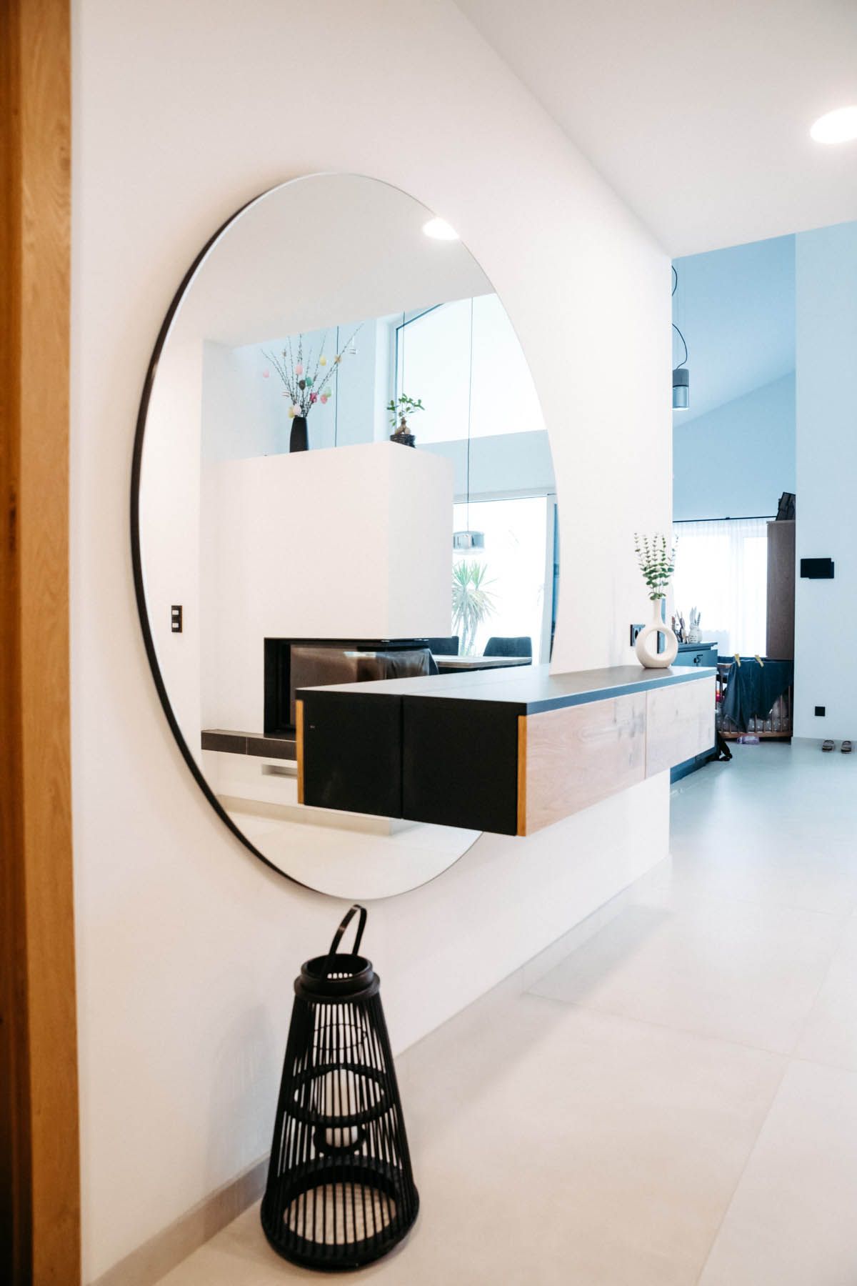 Großer runder Spiegel über einer schwebenden Holzkonsole. Eine schwarze Laterne steht auf dem Boden in einem weißen Flur.