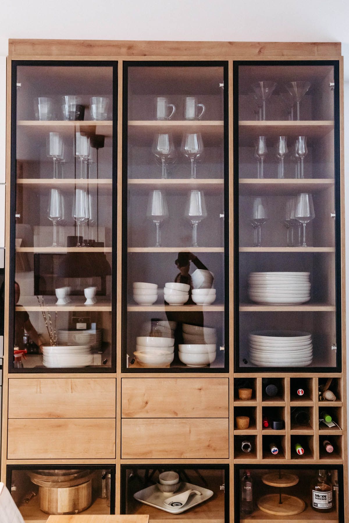 Holzschrank mit Glasfront, gefüllt mit Geschirr, Gläsern und Weinflaschen, in einer hellen Küche.