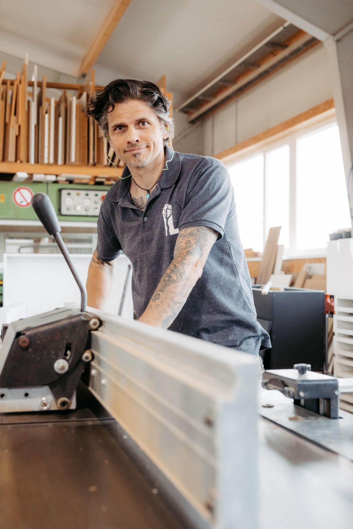 Ein lächelnder Tischler mit Armtattoos beugt sich in einer Werkstatt über eine Tischkreissäge.
