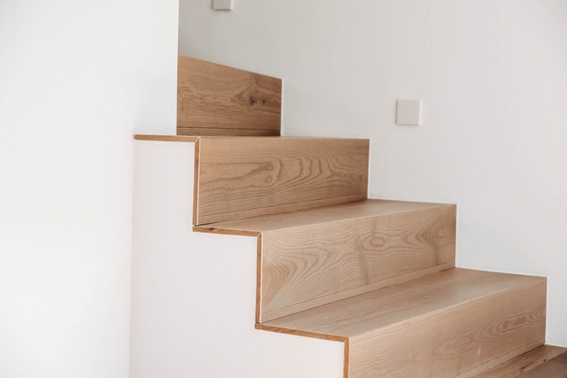 Holztreppe mit weißen Setzstufen vor einer weißen Wand.
