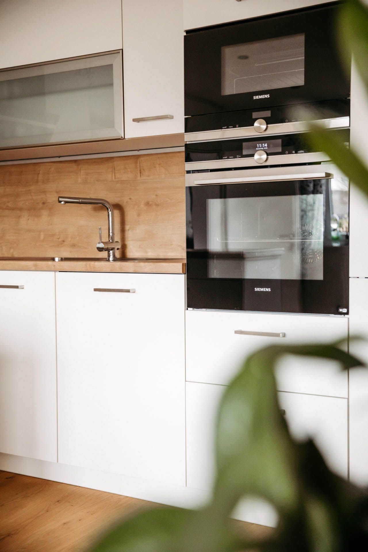 Moderne Küche mit weißen Schränken, Holzakzenten und schwarzem Ofen. Im Vordergrund steht eine Pflanze.