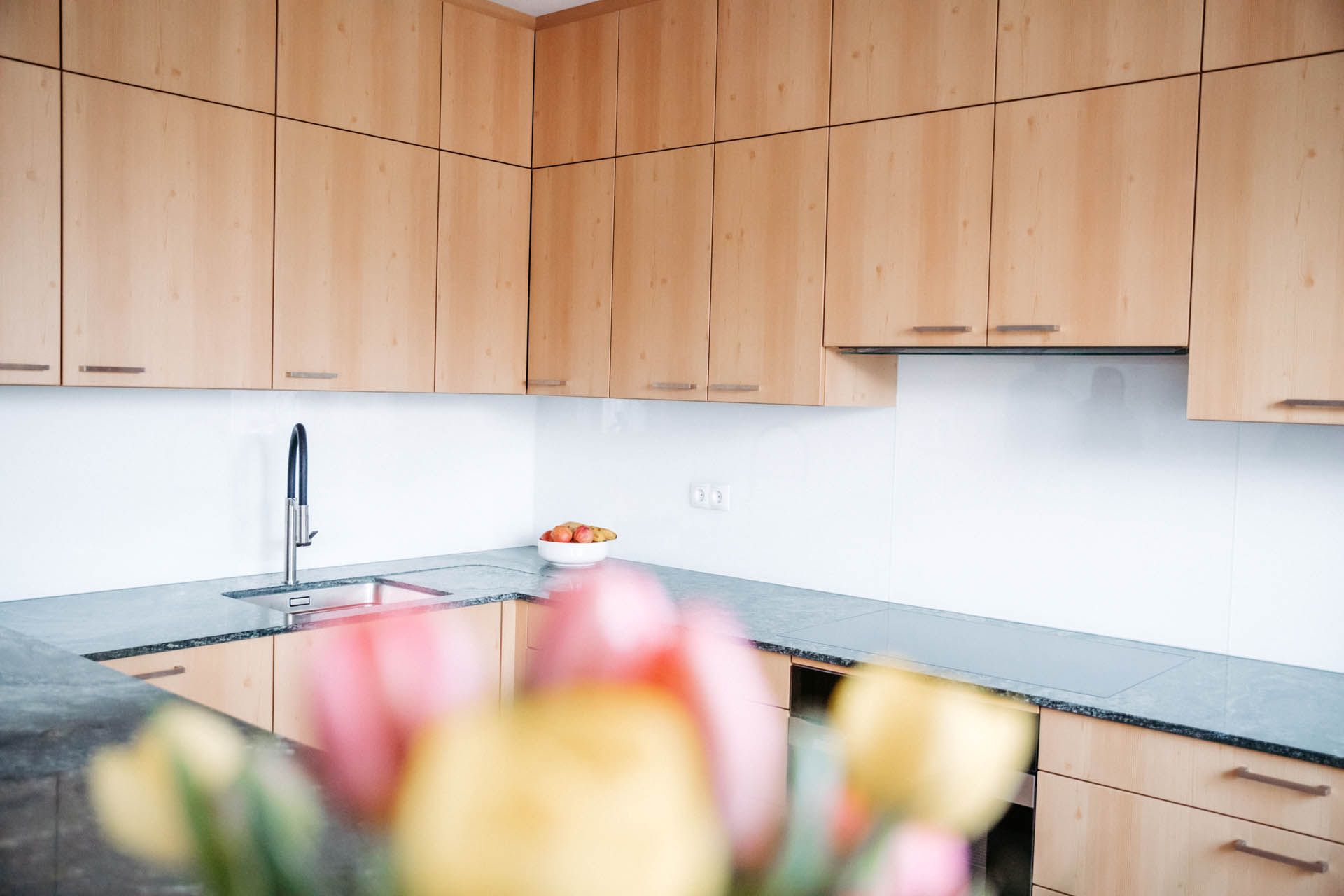 Küche mit hellen Holzschränken, dunklen Arbeitsplatten und einer Spüle. Im Vordergrund steht ein Blumenstrauß.