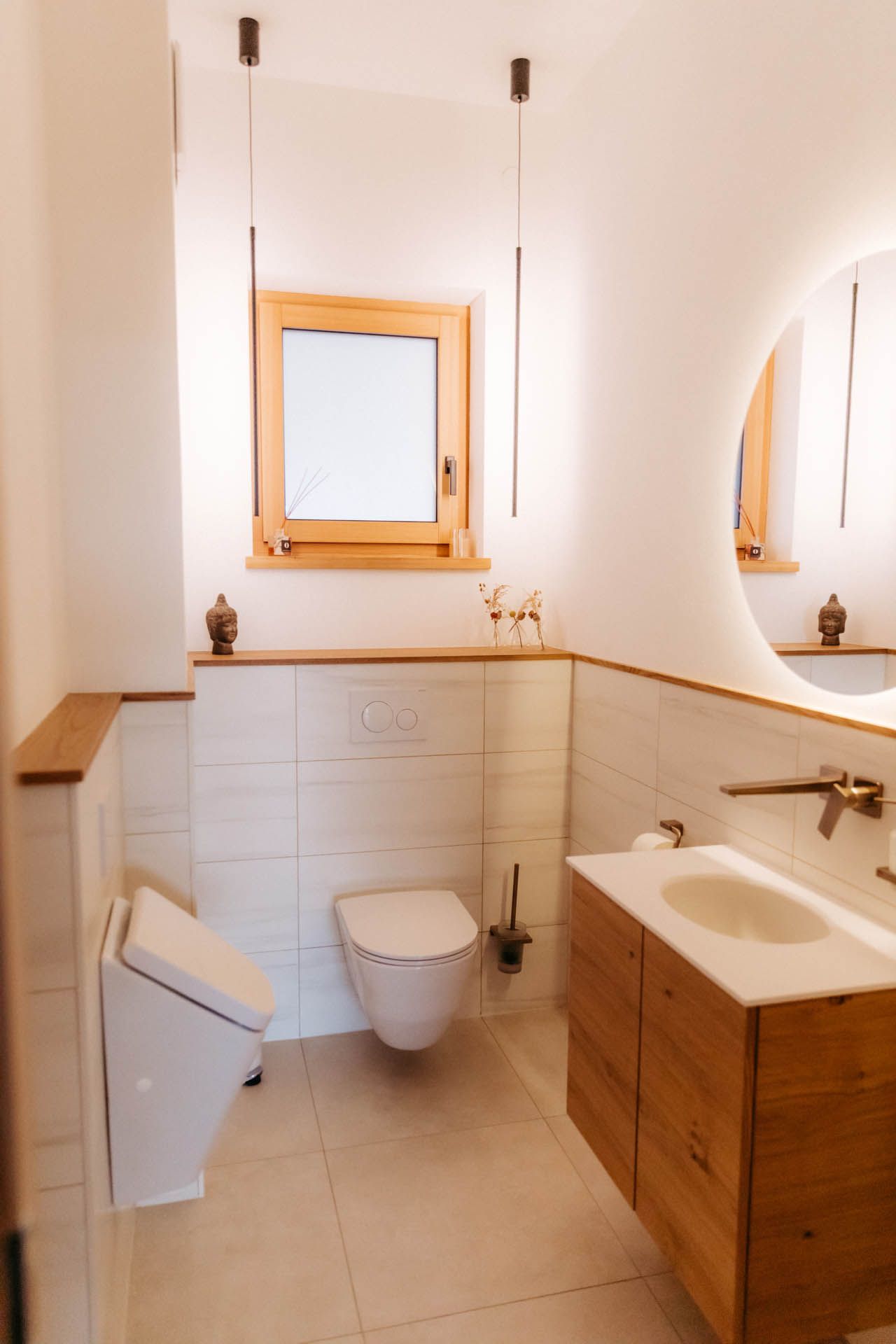 Modernes Badezimmer mit weißen Wänden, Holzakzenten und einem runden Spiegel. Toilette und Waschbecken sichtbar.