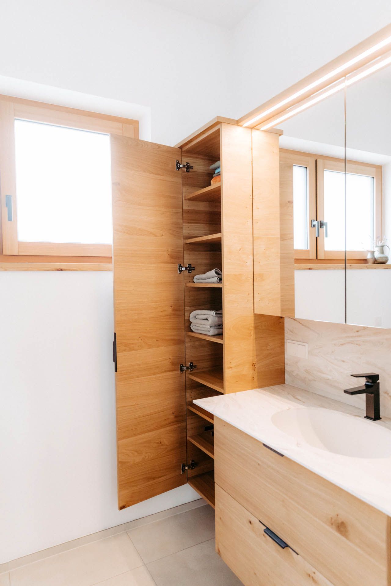 Badezimmer mit hölzernem Aufbewahrungsschrank für Handtücher; daneben ein Waschbecken und ein Spiegel.