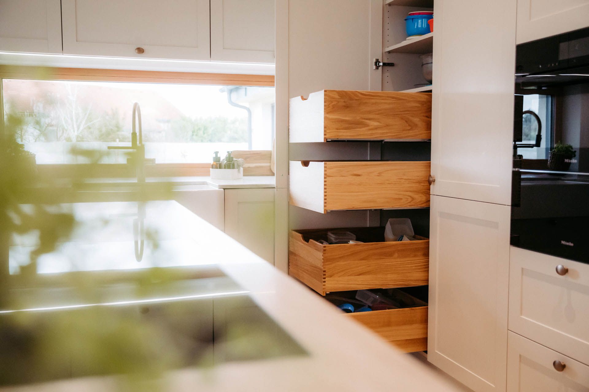 Weiße Küche mit offenen Holzschubladen, hinter denen Geschirr liegt, neben einem schwarzen Ofen und einer Spüle.