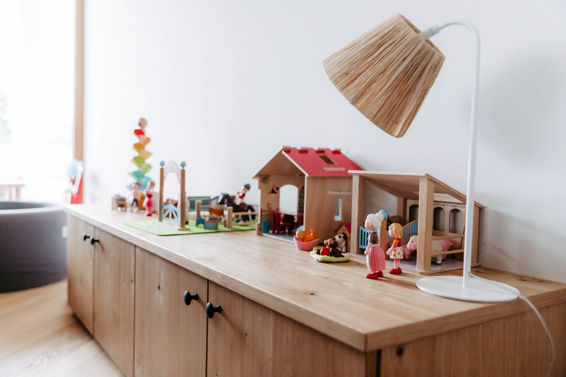 Holzspielzeughäuser und -figuren auf einem hellen Holzschrank mit einer Lampe vor einer weißen Wand.