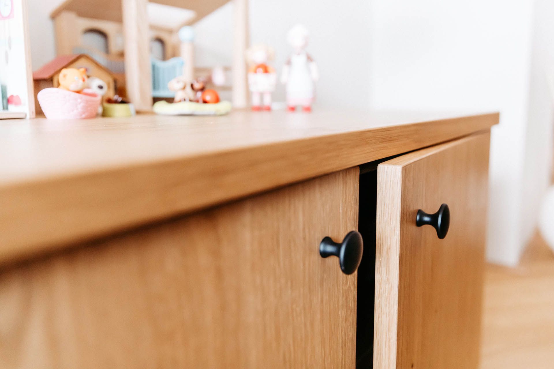 Holzschrank mit schwarzen Knöpfen, offene Tür gibt den Blick frei auf das Innere. Spielzeug liegt obenauf, in einem hellen Raum.