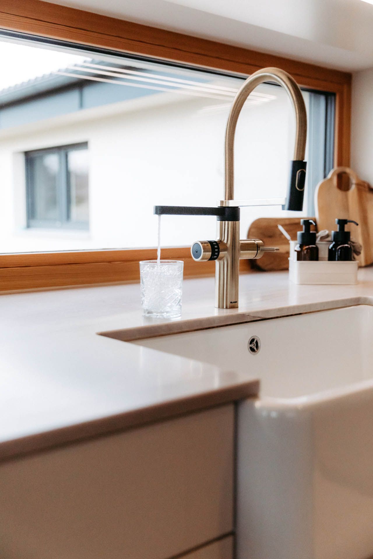 Goldener Küchenhahn, der ein Glas mit Wasser füllt und in einer weißen Spüle neben einem Fenster steht.