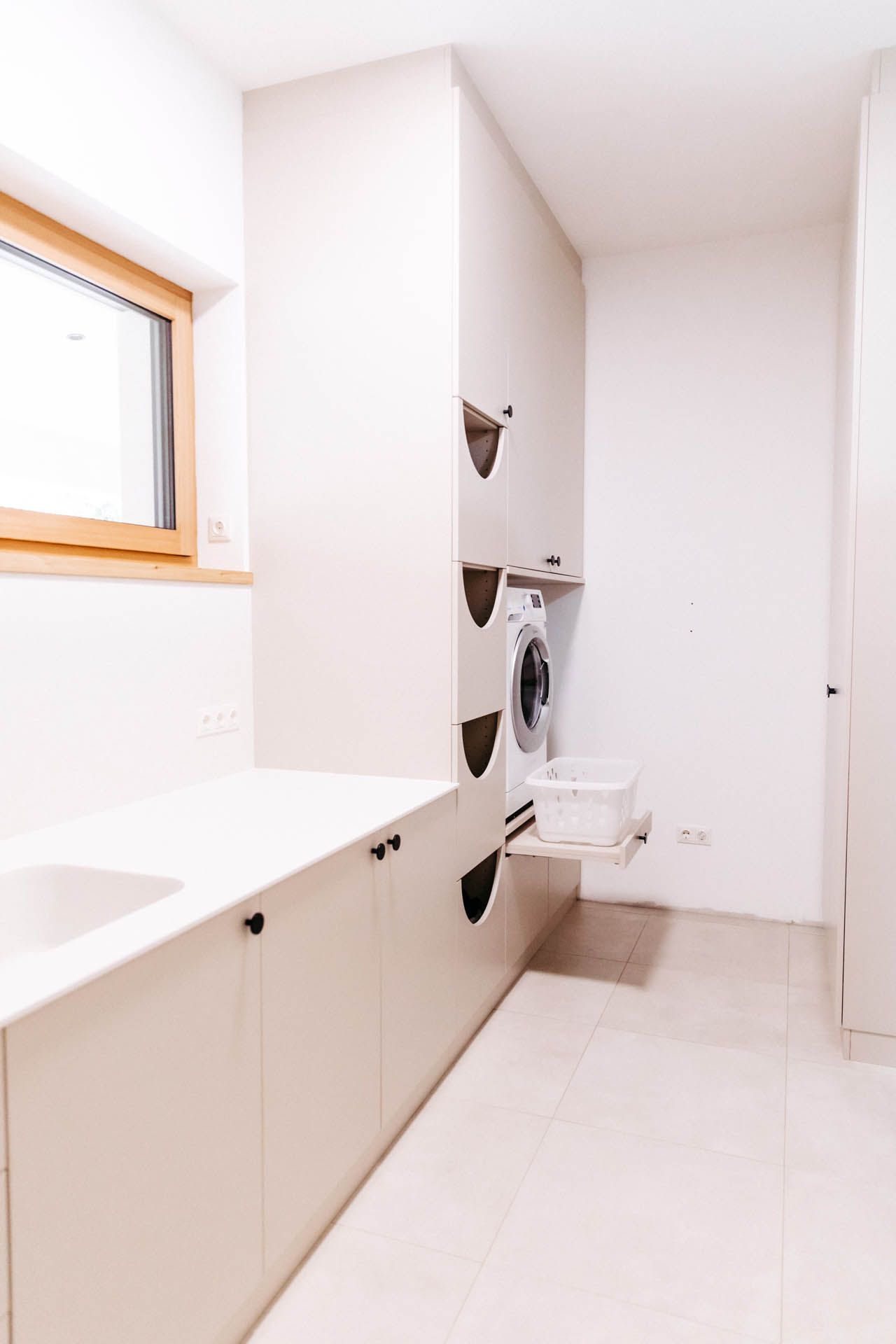 Waschküche mit weißen Schränken, Waschmaschine und ausziehbarem Wäschekorb; links ist ein Fenster.