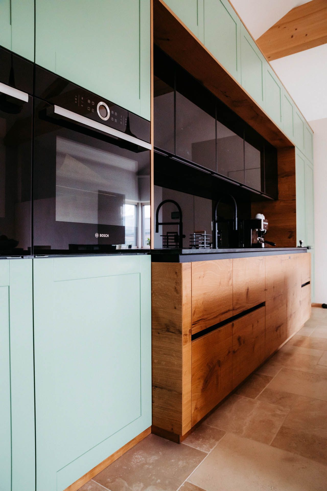 Moderne Küche mit mintgrünen Schränken, Holzinsel, schwarzen Geräten und Steinboden.
