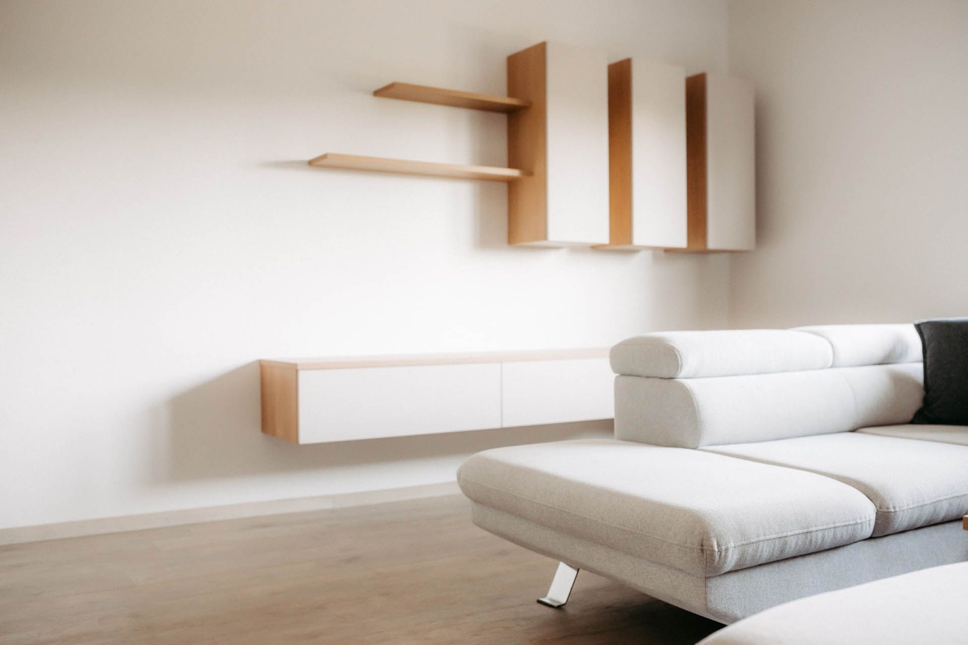Modernes Einbauregal und Schränke in Weiß und Holz an einer weißen Wand über einem hellgrauen Sofa.