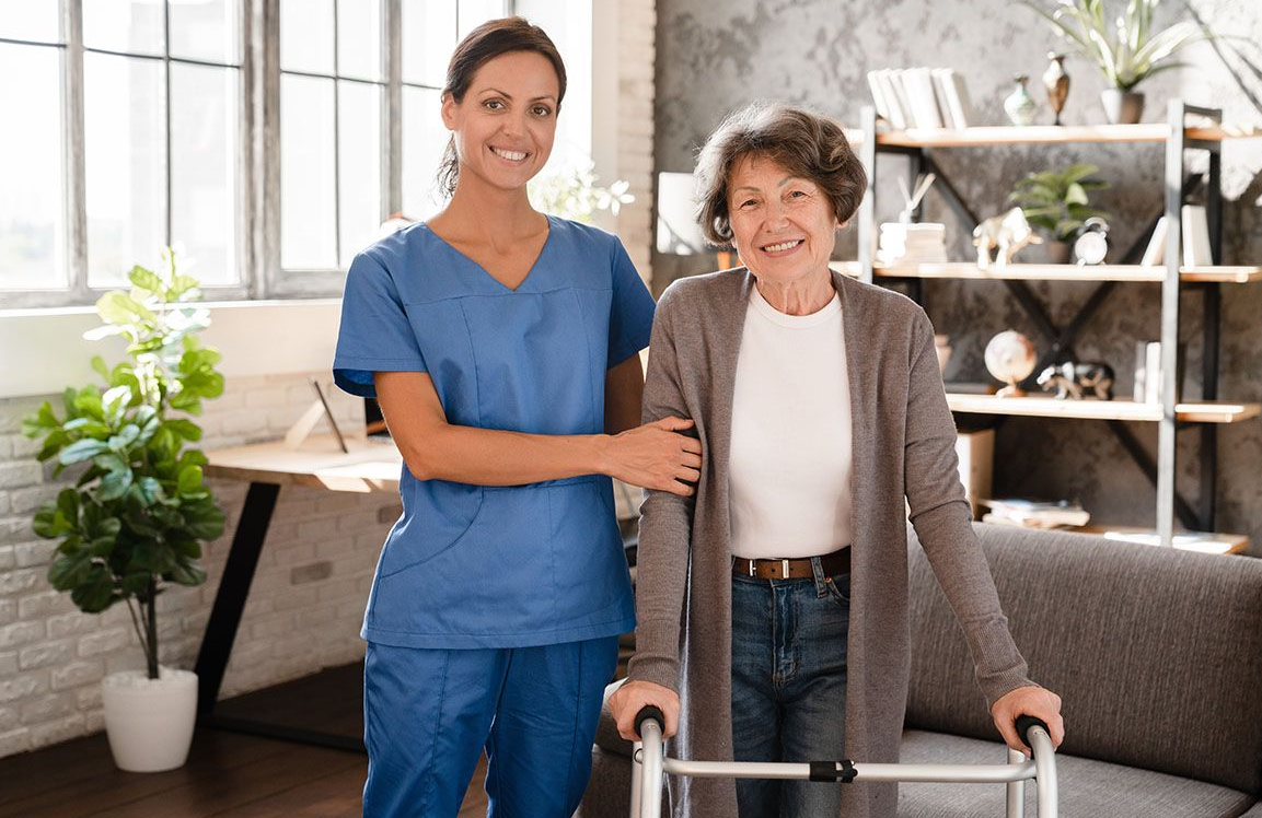 Caregiver assists older adult using a walker indoors. Both smiling. Blue scrubs, brown cardigan.