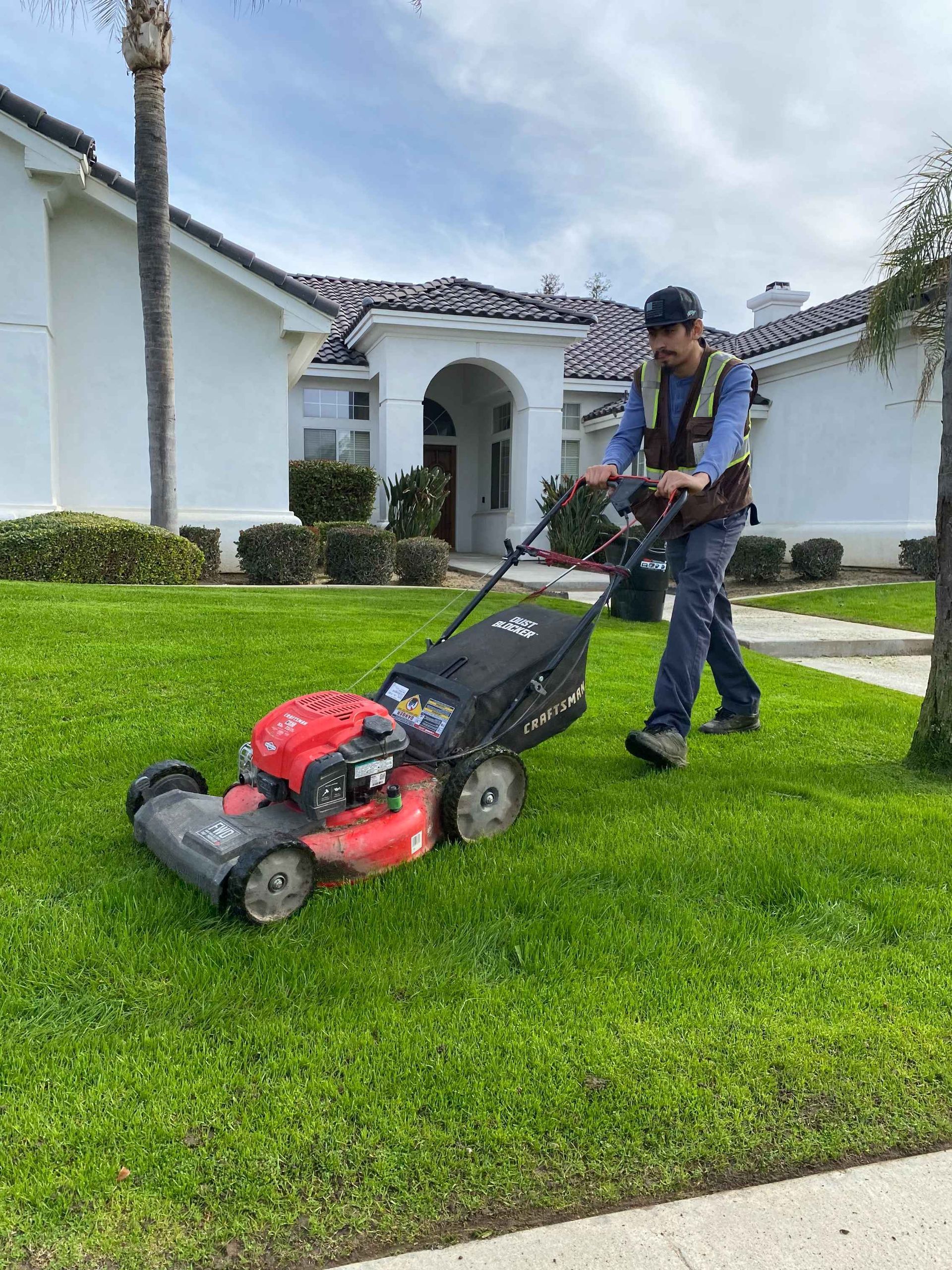 Man mowing a green lawn in front of a white house with a red lawnmower on a sunny day.