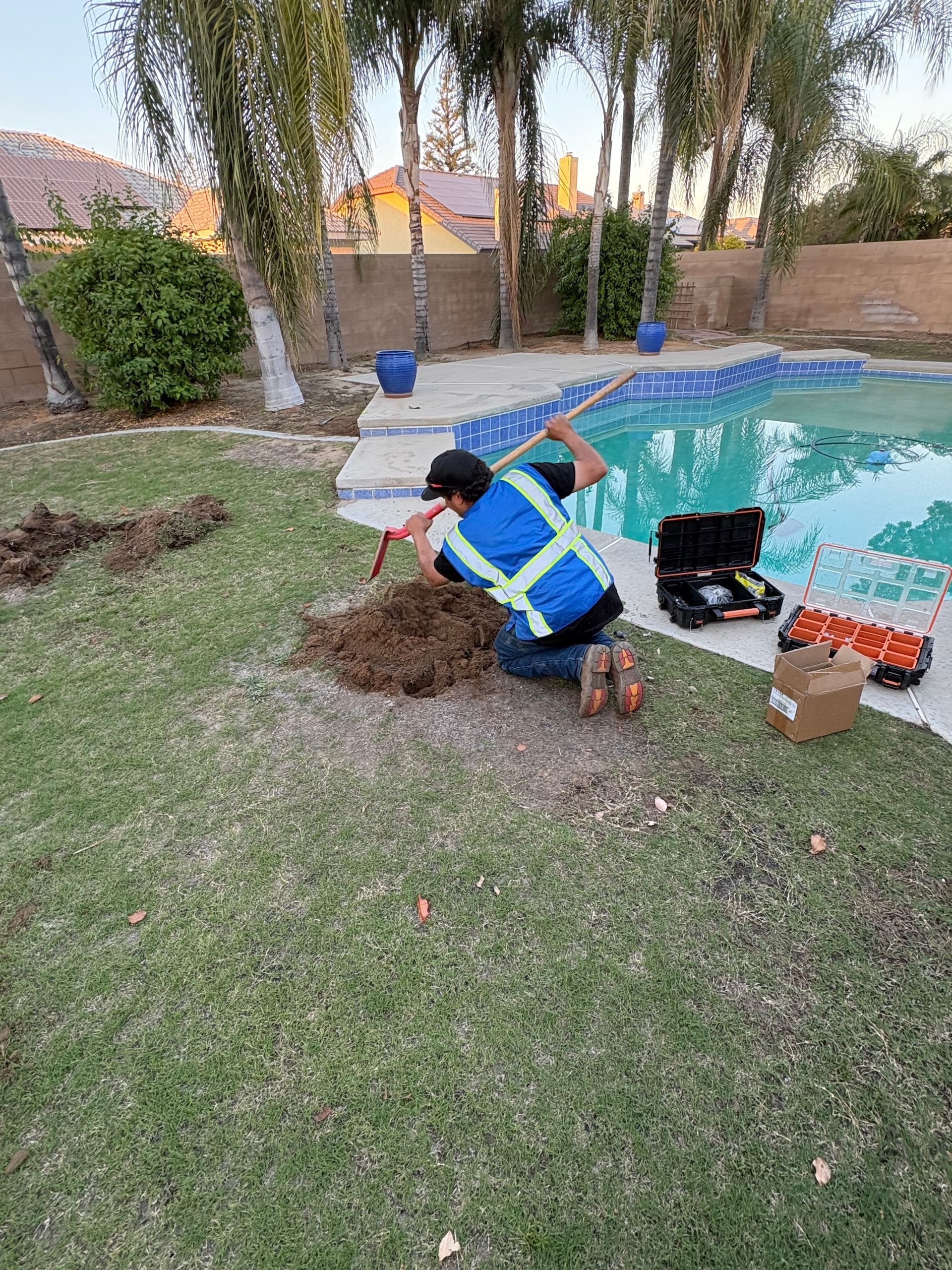 Man digging in yard near pool with tools and a reflective vest.
