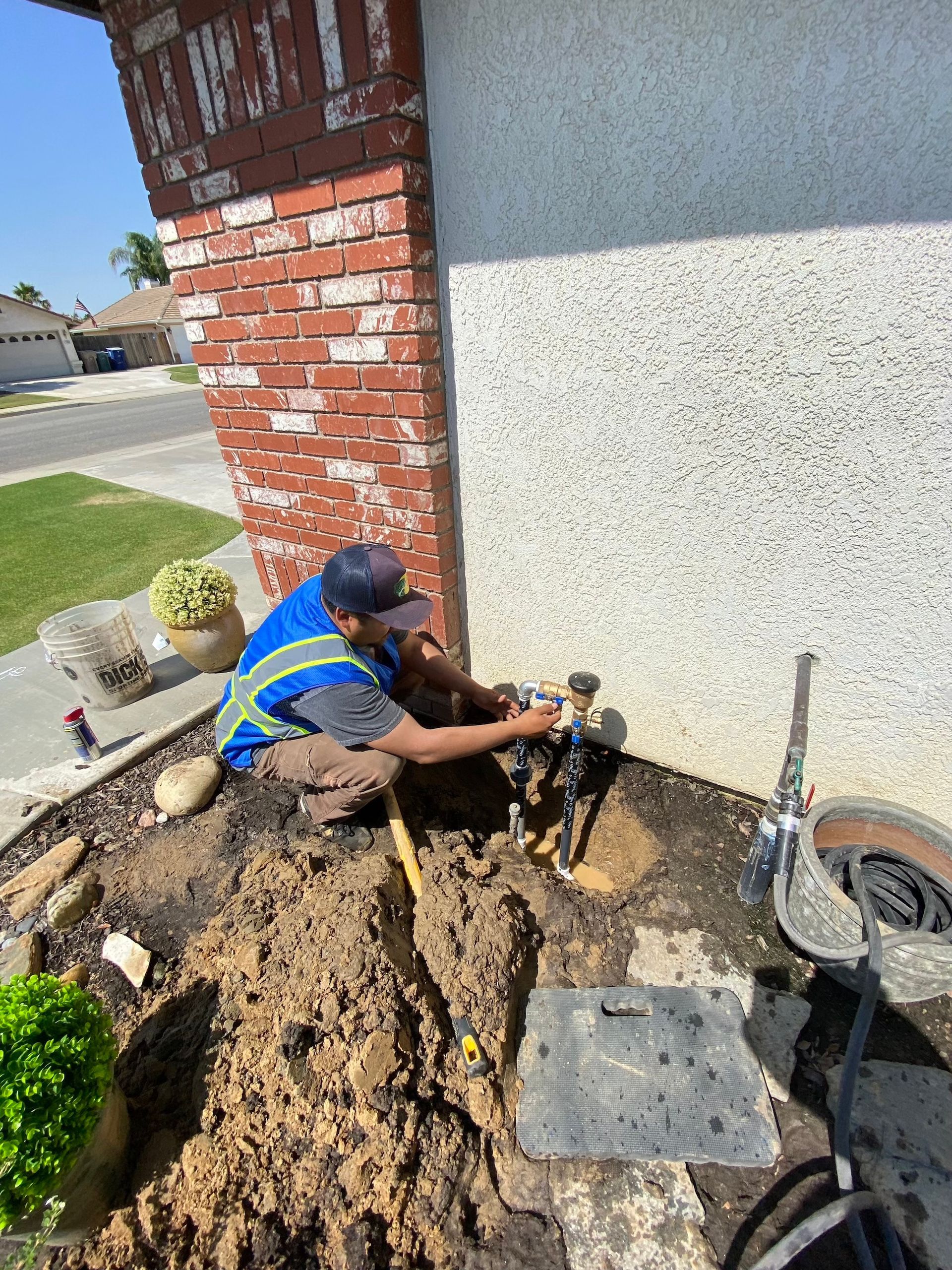 Person installing sprinkler system near a brick pillar and house, with exposed dirt and tools.