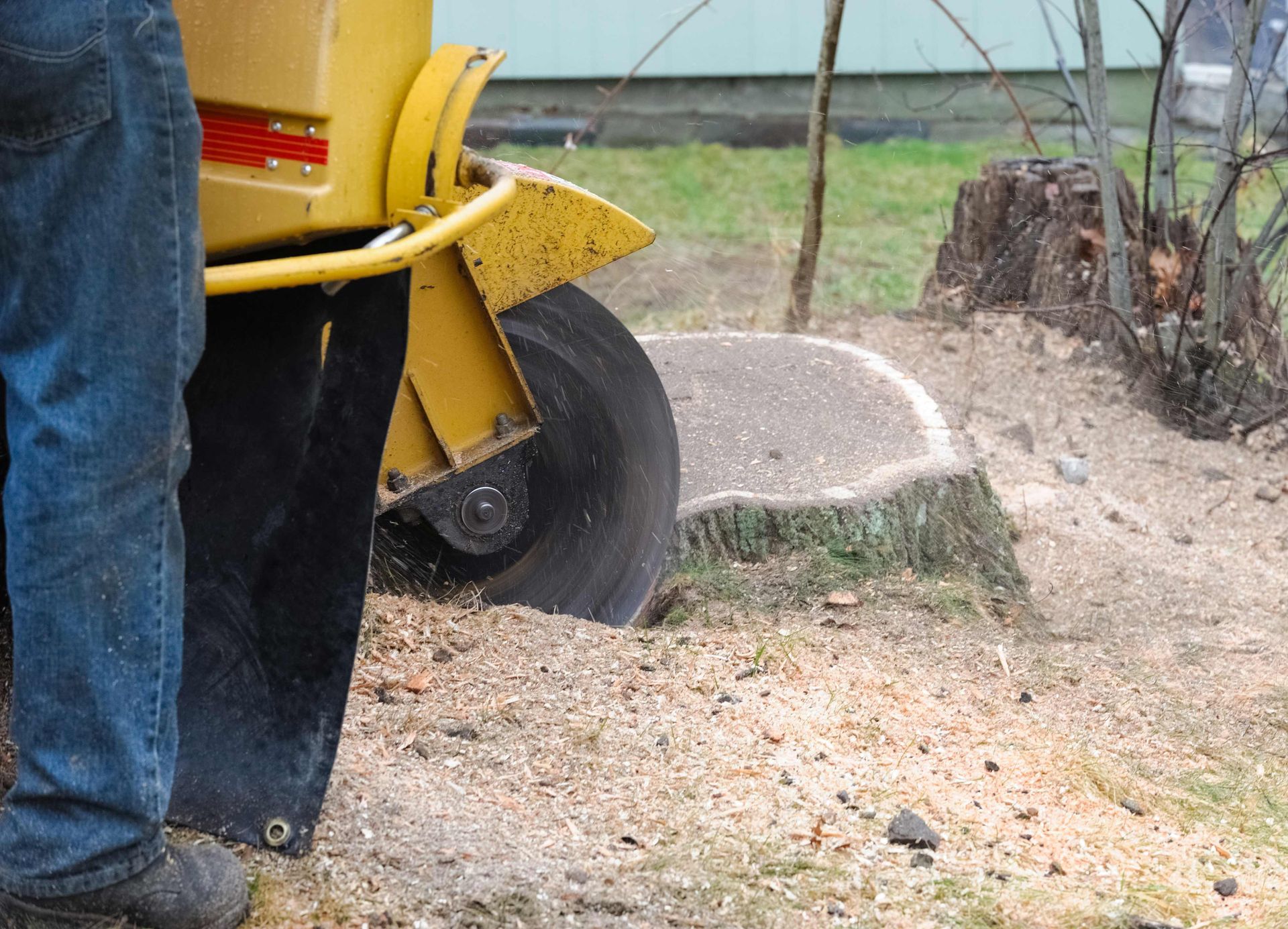 A yellow wood chipper cuts tree branches beside a person’s jeans-clad leg on a dirt path.