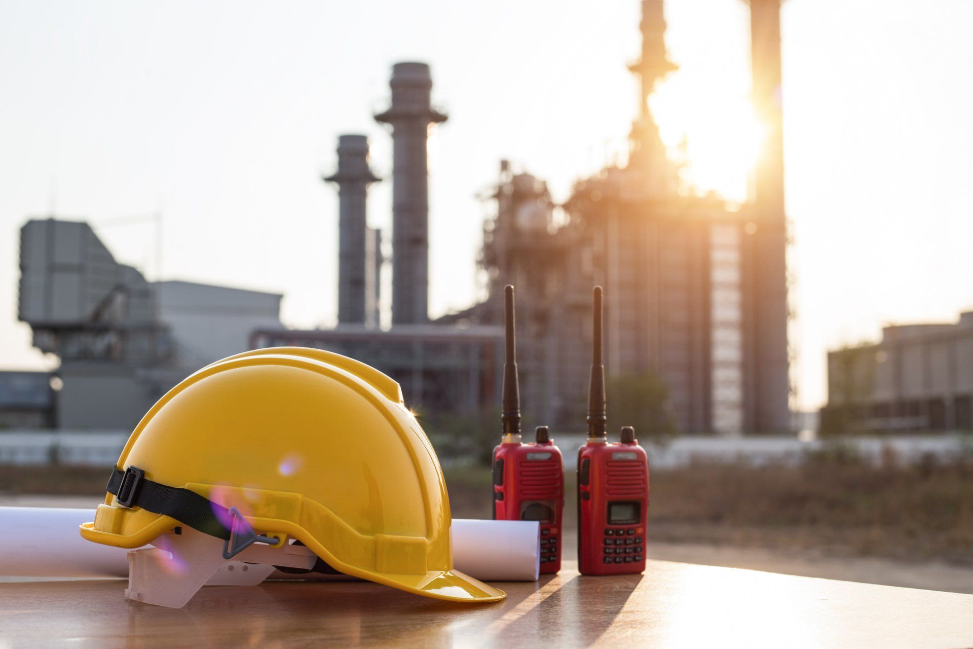 Yellow hard hat and two-way radios on a table in front of a power plant in sunlight.