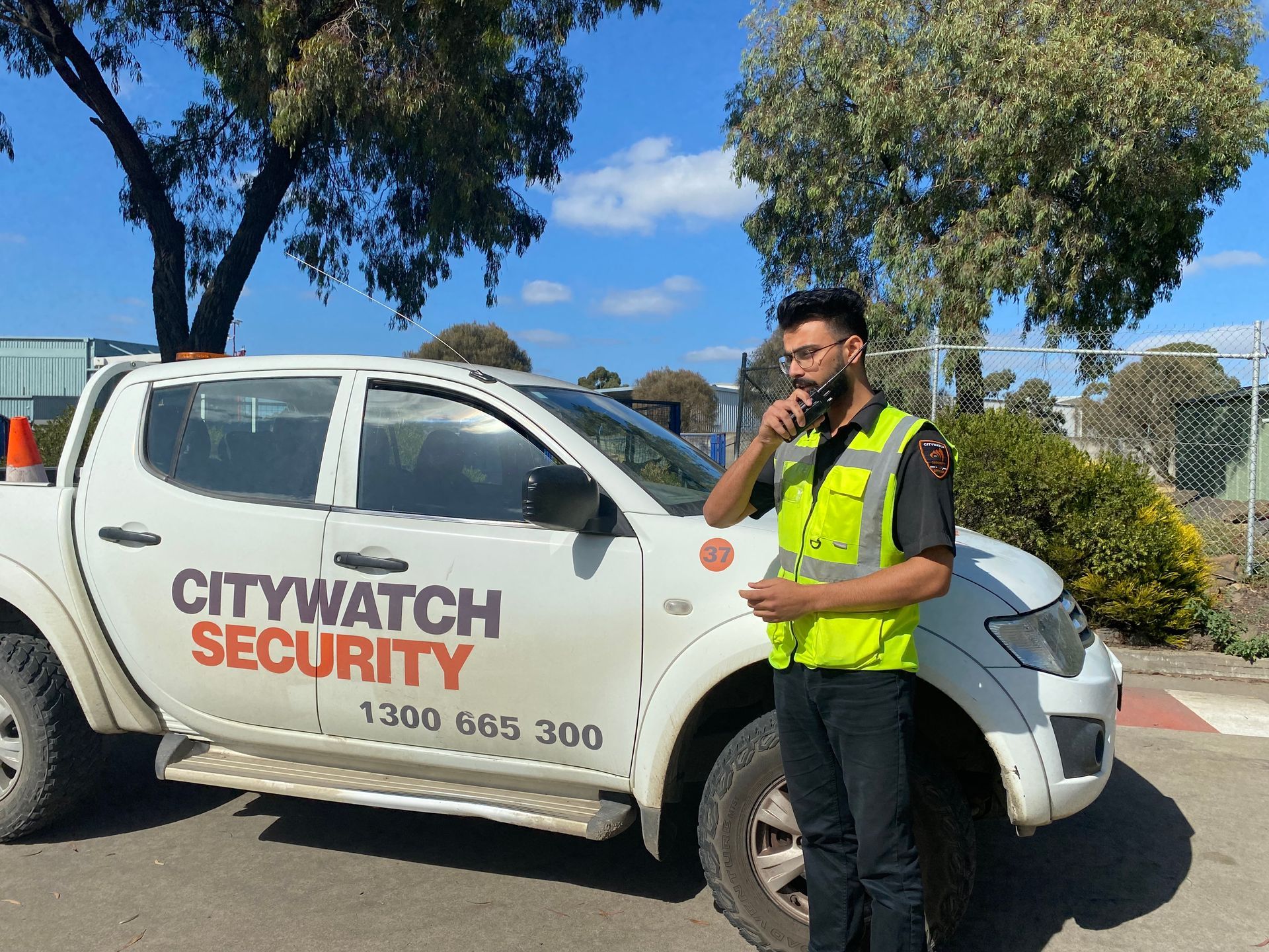 Security guard in a yellow vest stands by a Citywatch Security truck, under a blue sky.