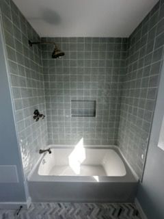 A small, gray tiled shower with an alcove, bathtub, and bronze showerhead. Bright light shines on the tub.