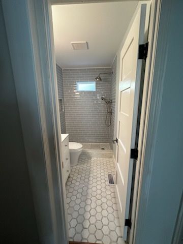 View of a small, renovated bathroom with hexagonal tile flooring, gray subway tile shower, and white door.