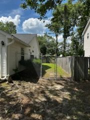Backyard view with a chain-link fence gate opening to a narrow grassy passage between wooden fences and a white house under a blue sky.
