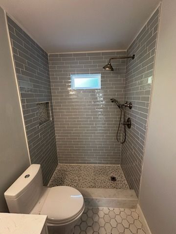 A small bathroom featuring a tiled shower and toilet. The shower has gray brick-style tiles with pebble flooring.