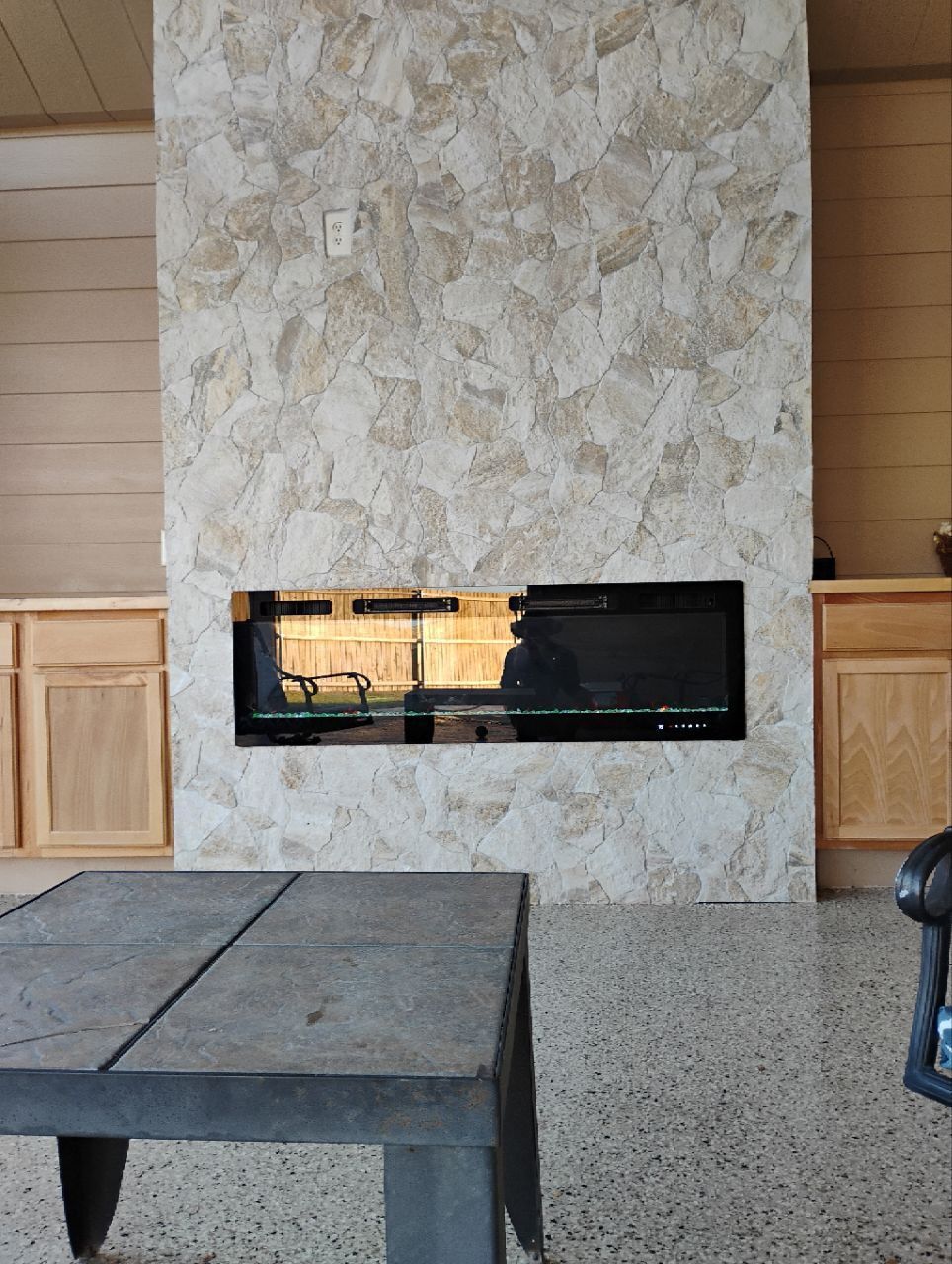 Fireplace centered on a stone-covered wall, flanked by wooden cabinets; a dark table in the foreground.