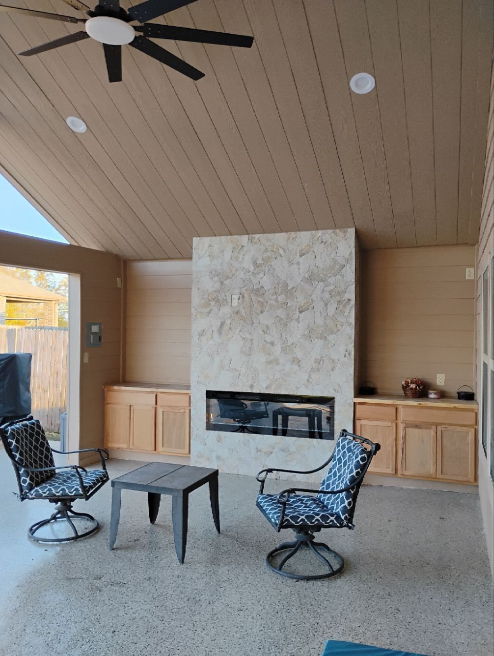 Outdoor patio with fireplace, seating, and a ceiling fan. Beige, gray, and black colors dominate the space.