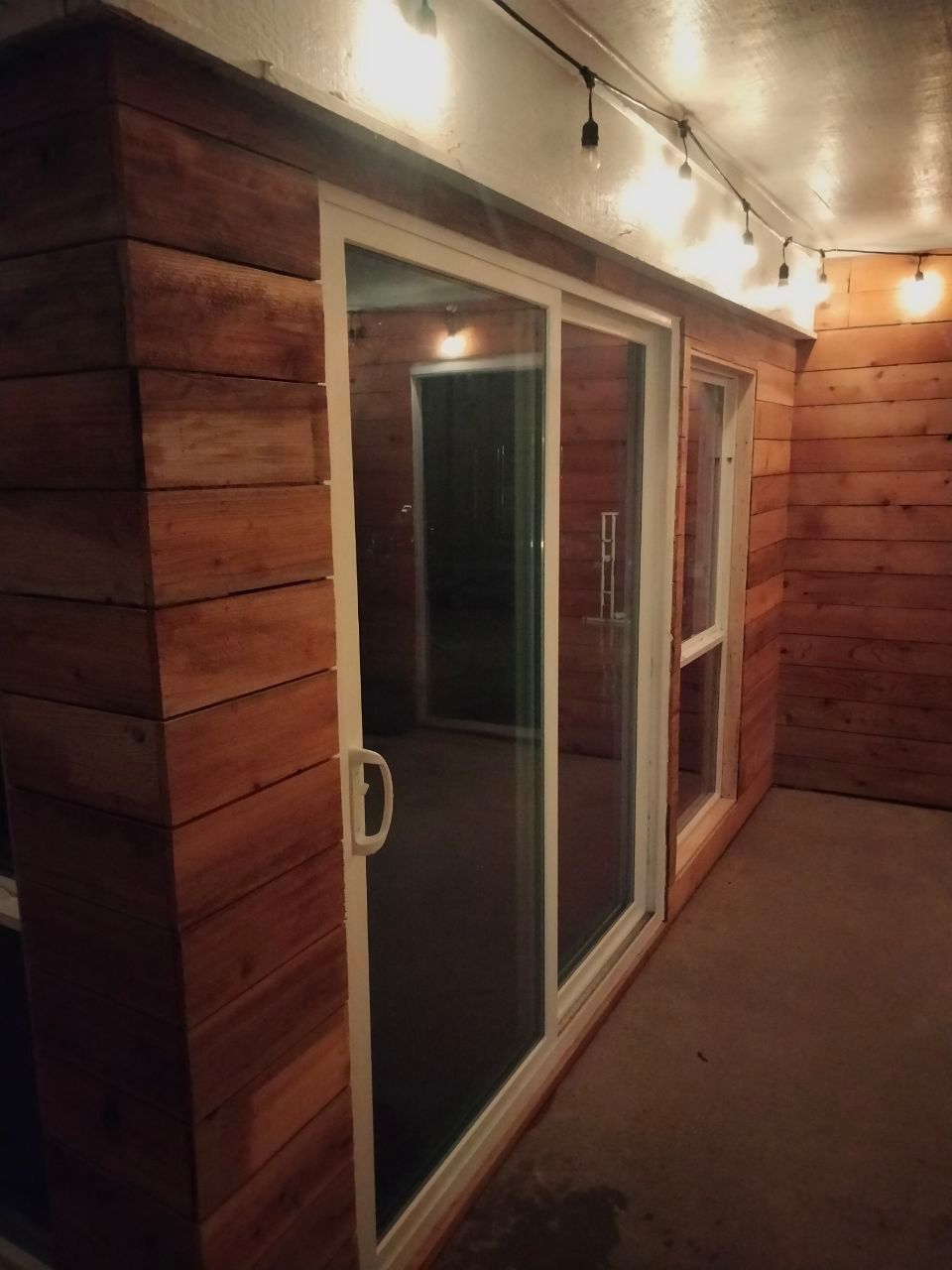 Wooden-paneled room with white-framed sliding glass door and small window. String lights illuminate the space.