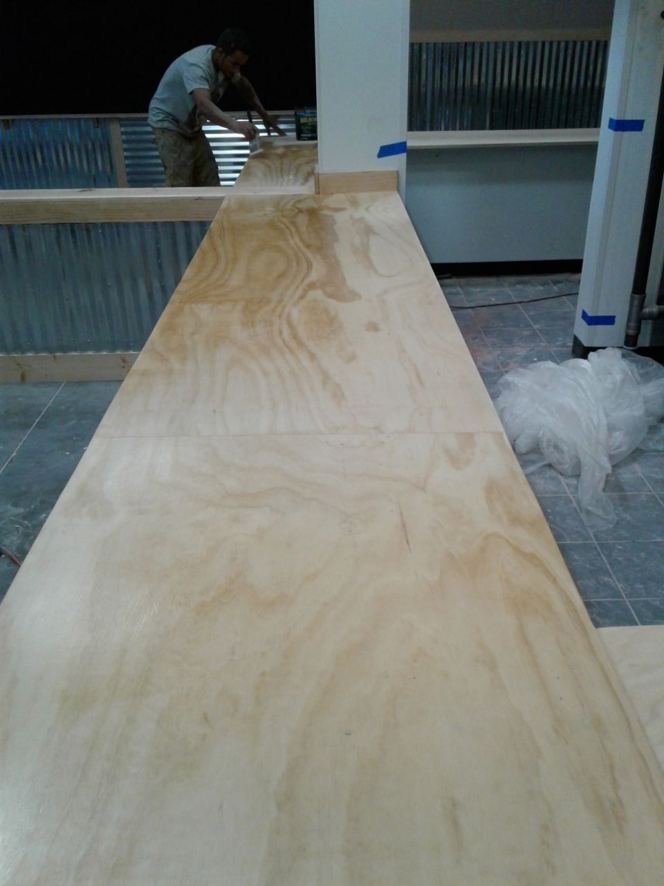 Person sanding a long, light-wood counter. Corrugated metal and shelving in the background.