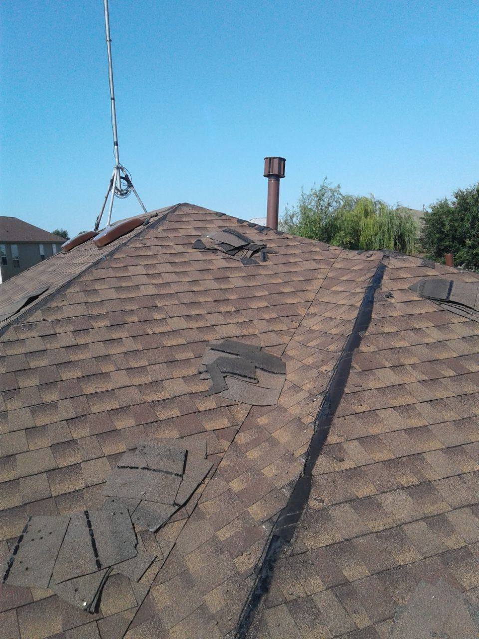 Damaged asphalt shingle roof, torn sections visible, with a chimney and antenna against a blue sky.