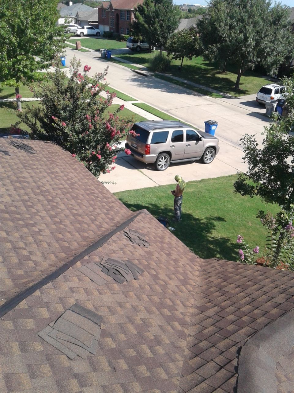Overhead view of a house roof with damage, looking out to a suburban street with a silver SUV parked.