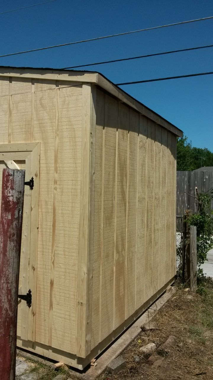 Side view of a light brown shed with vertical siding, black hardware, and a dark roof under a blue sky.