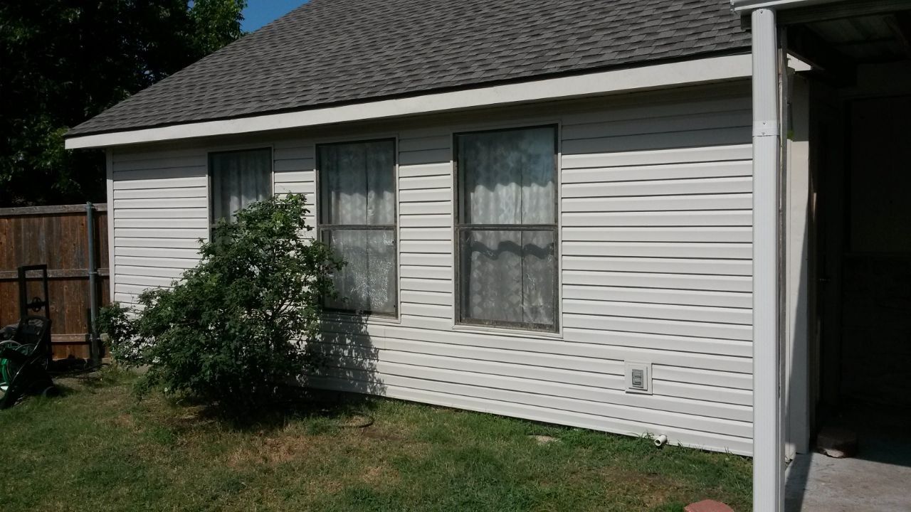 A small house with white siding and a gray roof, with three windows and a bush in front.