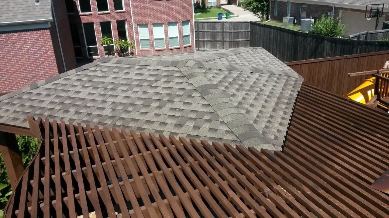 Brown wooden pergola with a gray shingled roof, next to a brick building and a wooden fence.