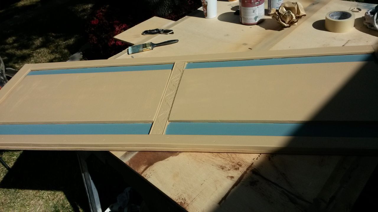 A beige painted wooden door with blue trim is on a work surface, painting supplies nearby.