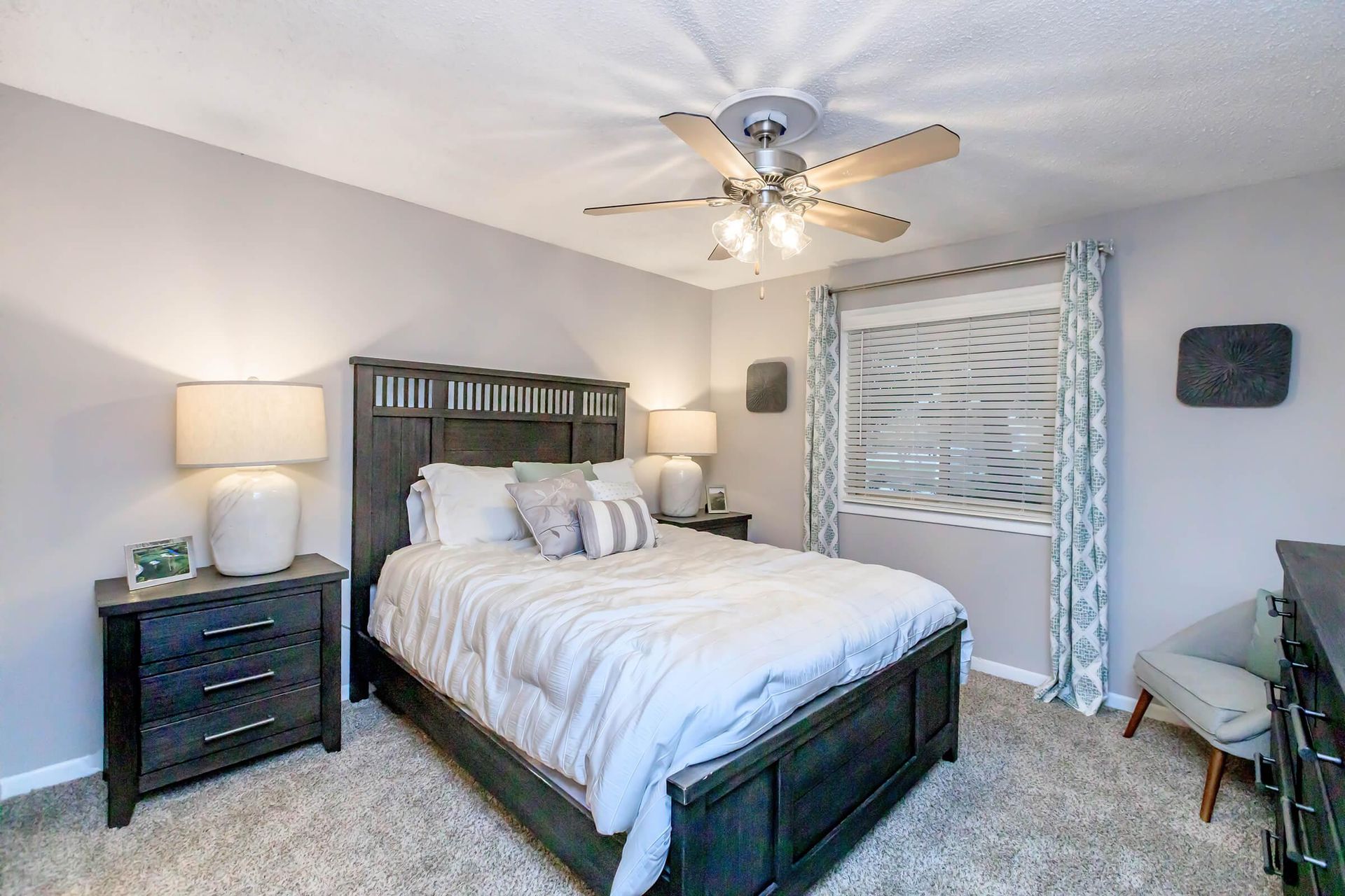 Bedroom with a bed, nightstands, a ceiling fan, window, and a chair. Soft lighting and neutral colors.