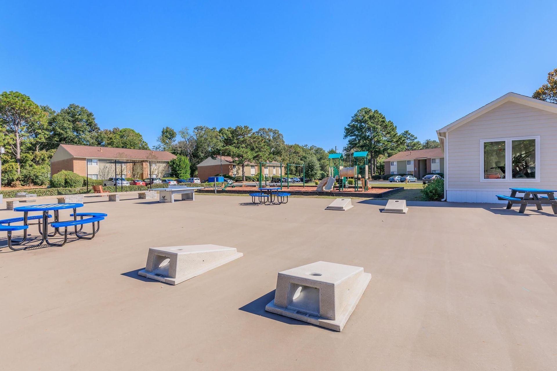 A concrete recreation area with picnic tables, cornhole boards, and a playground in front of buildings under a clear, blue sky.