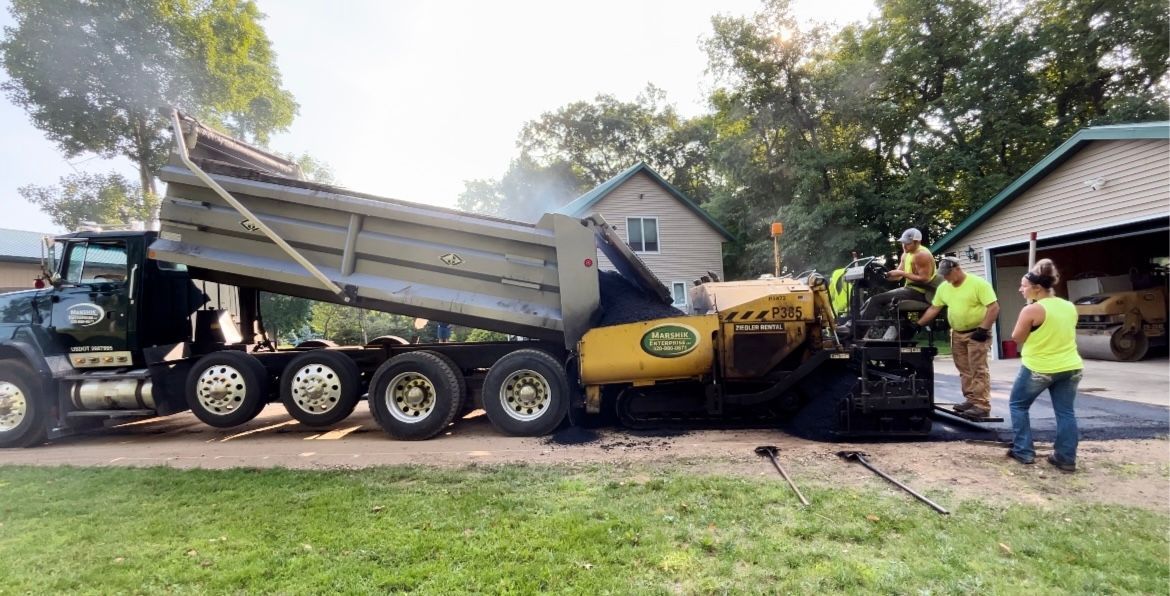 A dump truck is being loaded with asphalt in a driveway.