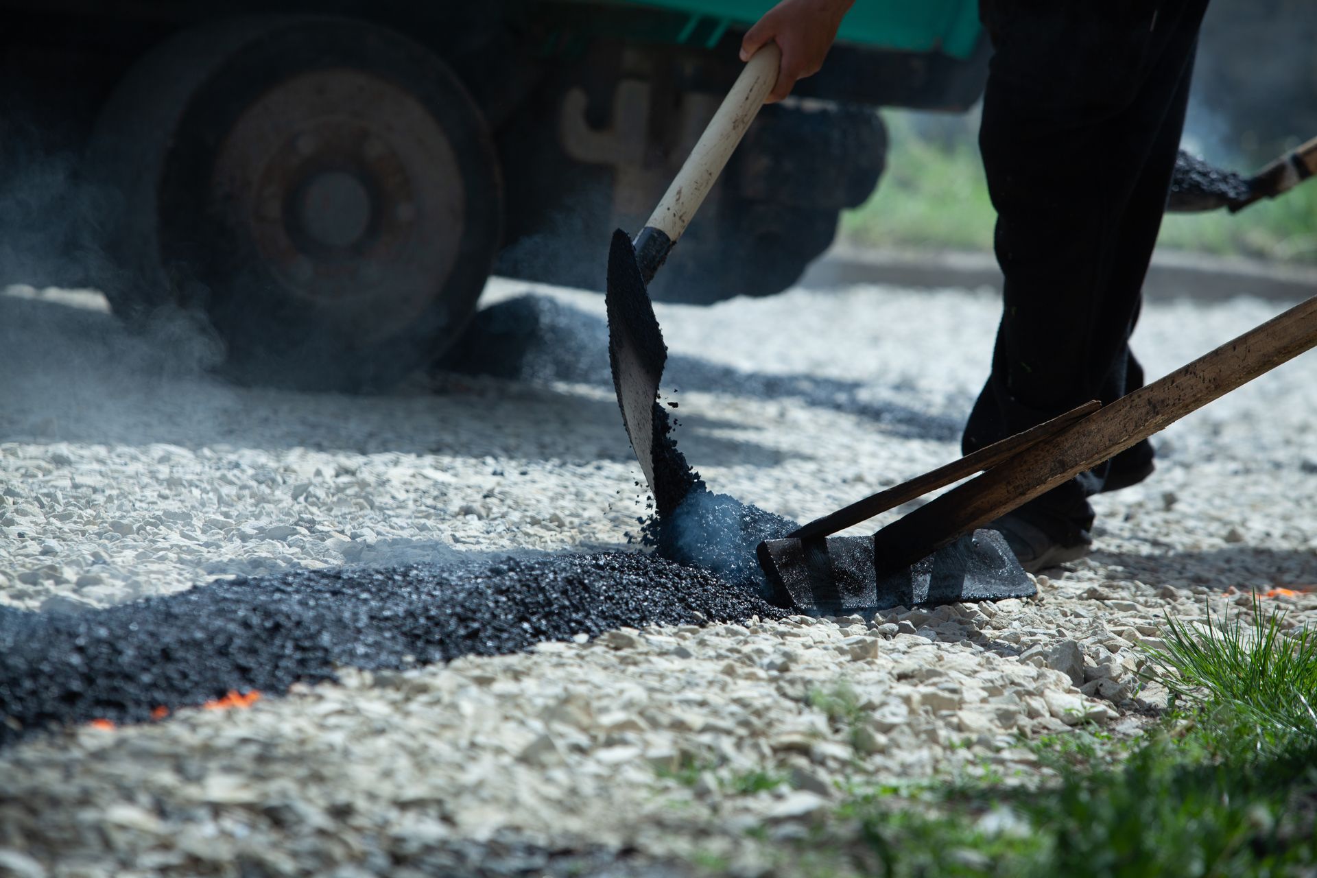 Person using tools to spread asphalt on a road. A vehicle is in the background.