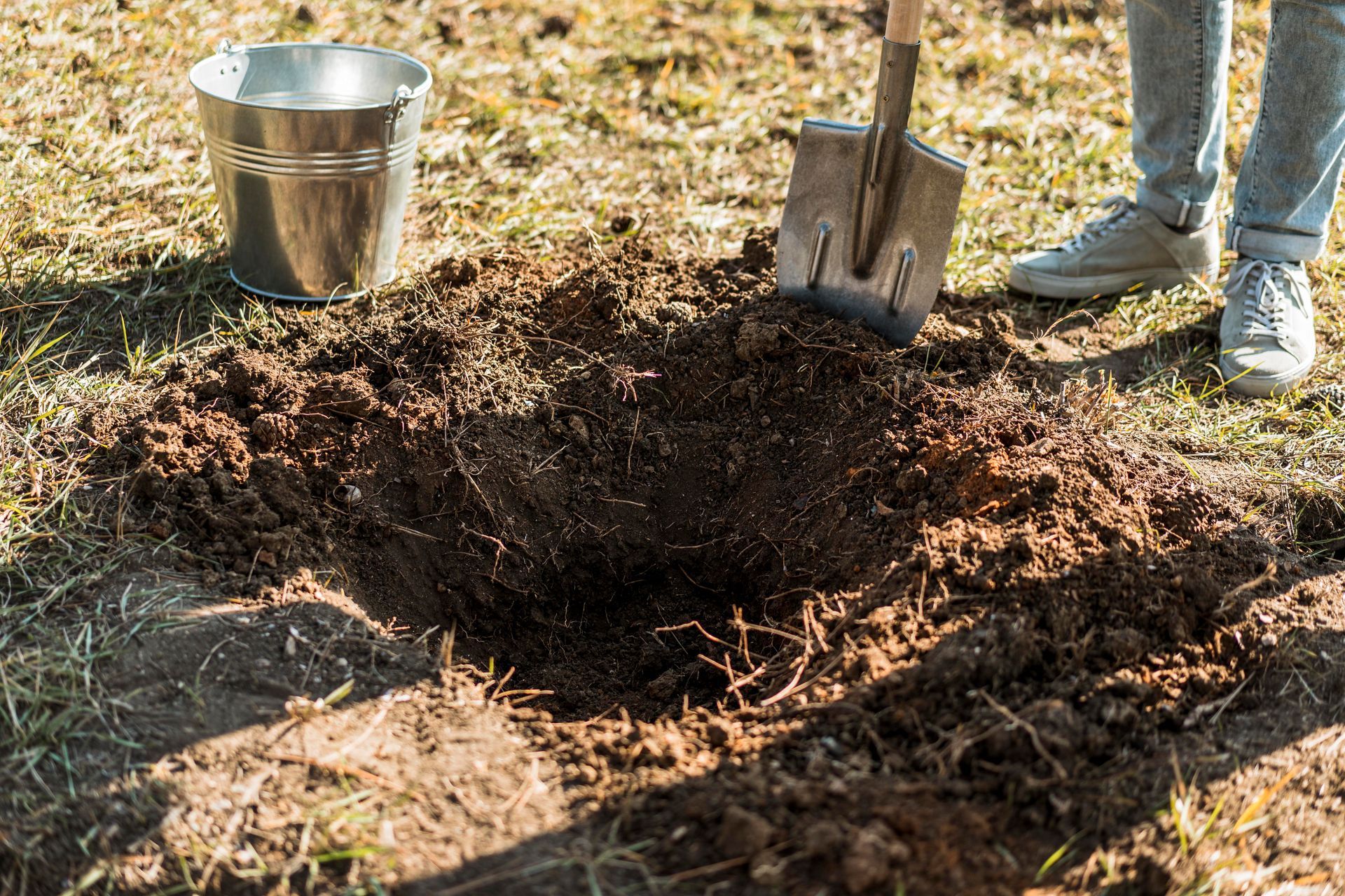 Person digging a hole in the ground with a shovel, with a bucket nearby.