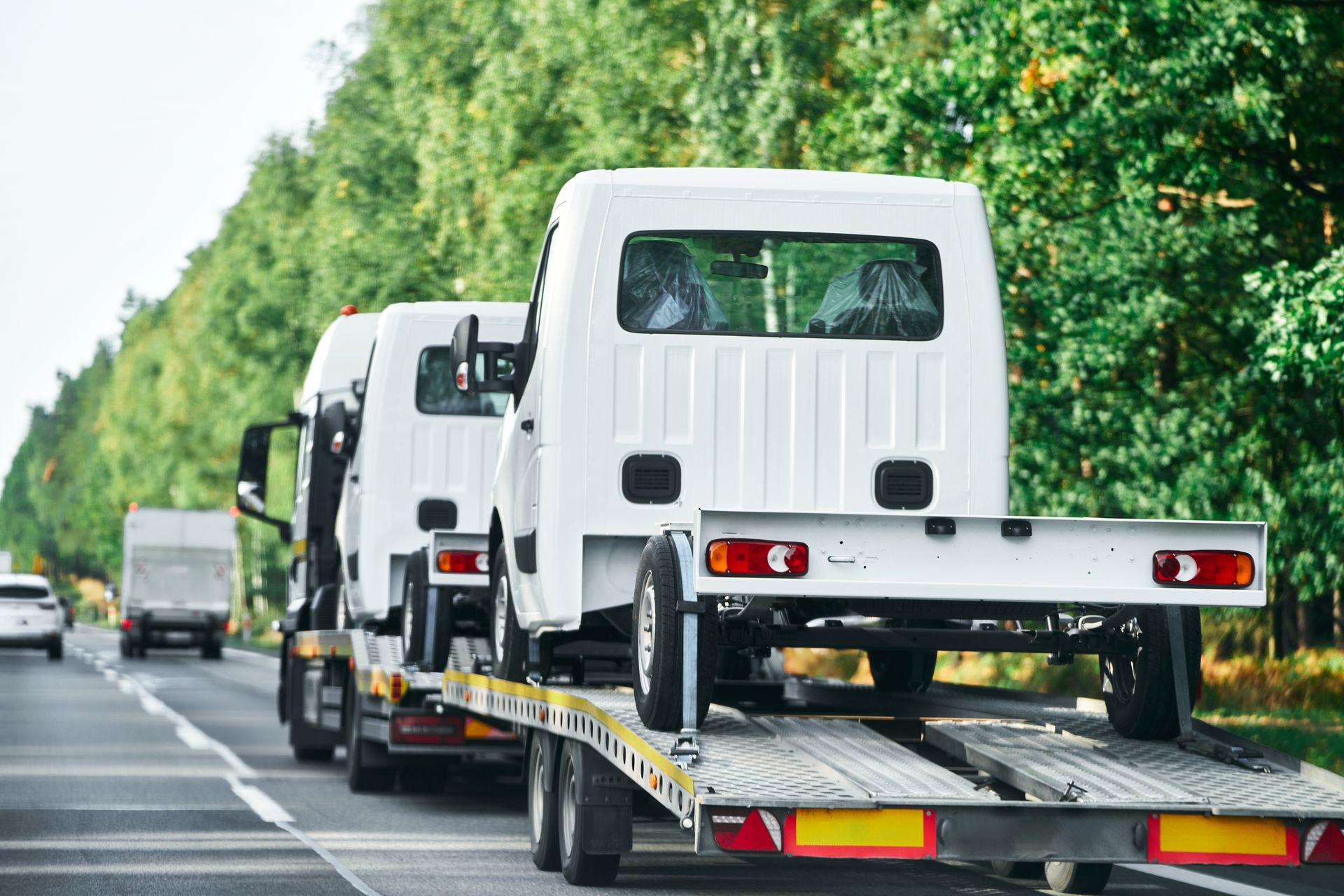 White trucks on a trailer traveling down a highway, trees in the background. — Geelong Accident Towing & Recovery Centre In North Geelong, VIC