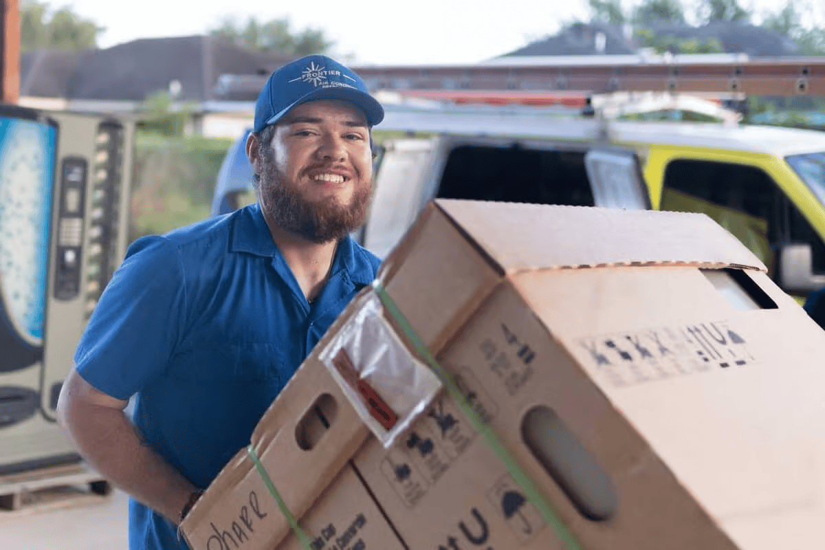 Smiling man in blue uniform carrying stacked cardboard boxes, near a van and a vending machine.