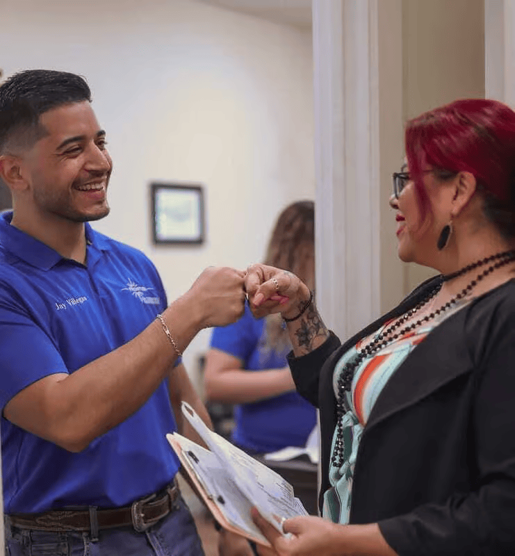Two people fist-bumping. Man in blue shirt smiles. Woman with red hair holds papers and smiles. Indoor setting.