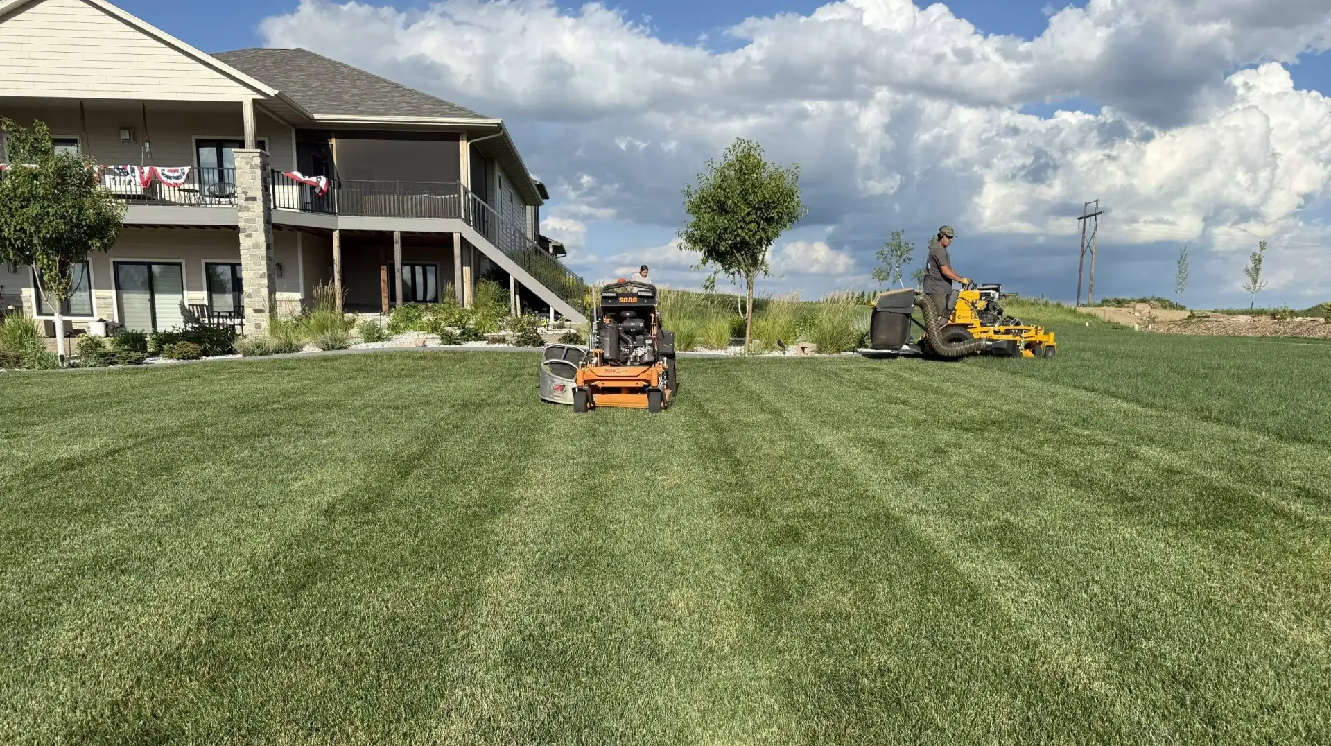 Two orange and yellow lawn mowers work on a large, freshly cut residential lawn near a two-story house.