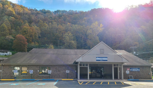 Brick building entrance with covered porch, parked car, and wooded hillside in the background