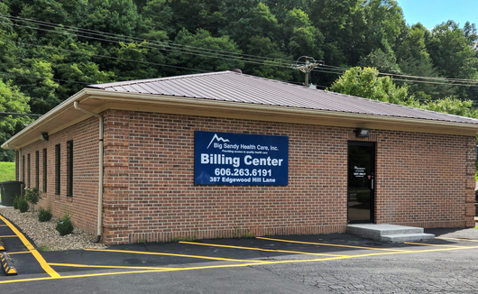 Brick billing center building with blue sign and black roof, surrounded by trees and yellow curb lines