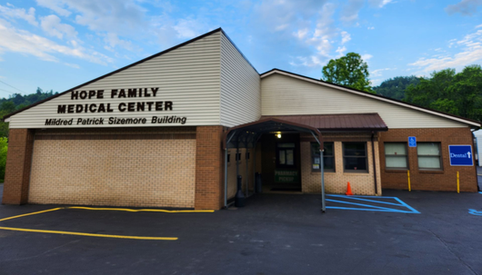 Brick building entrance with covered porch, parked car, and wooded hillside in the background