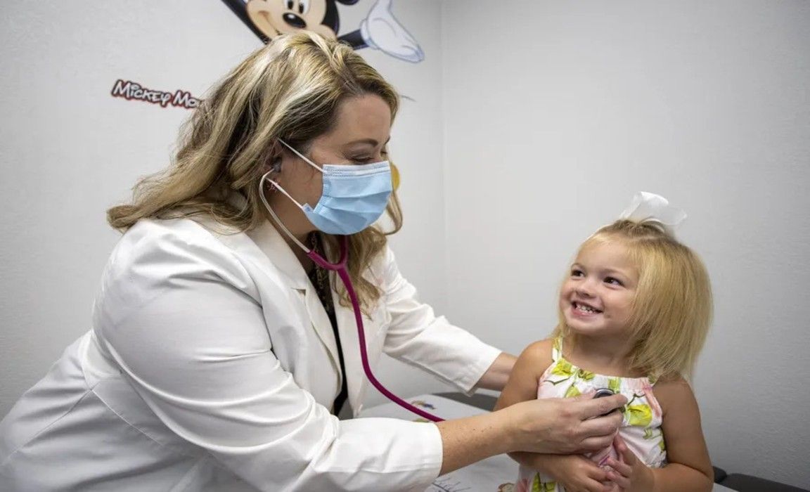 Doctor wearing a mask examines a smiling child in a clinic room.