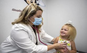 Doctor wearing a mask examines a smiling child in a clinic room.